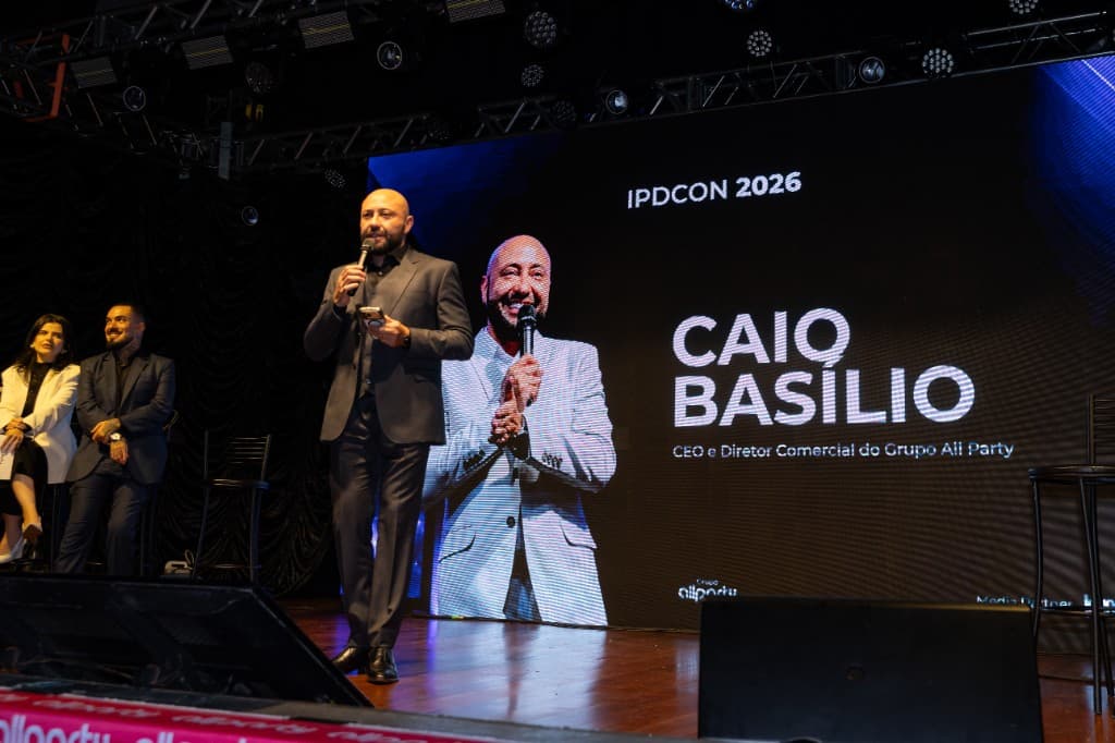 Caio Basílio no palco do IPDCON 2026, com o painel de LED anunciando seu nome